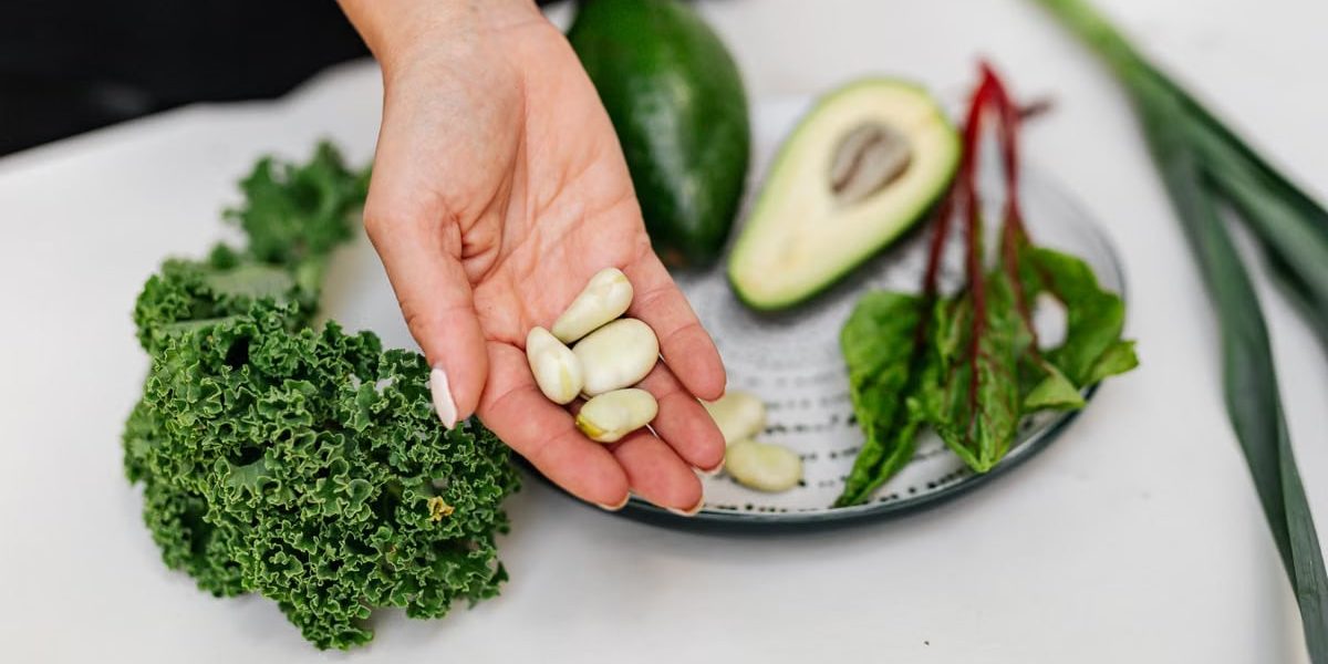 Close-up of a hand holding garlic cloves over a plate with fresh vegetables, including kale and avocado.