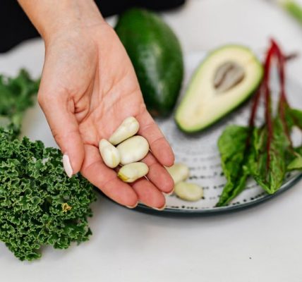 Close-up of a hand holding garlic cloves over a plate with fresh vegetables, including kale and avocado.