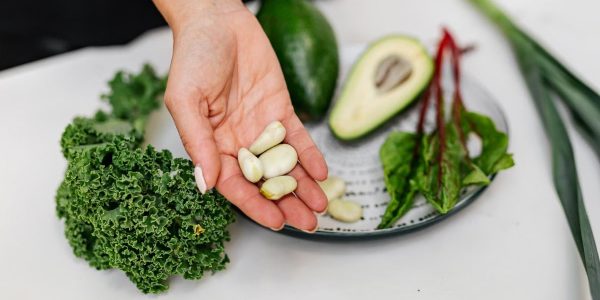Close-up of a hand holding garlic cloves over a plate with fresh vegetables, including kale and avocado.