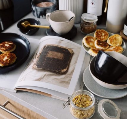 High angle of cookbook and cooked fritters on plates placed on table near utensils and ingredients in cozy kitchen at home