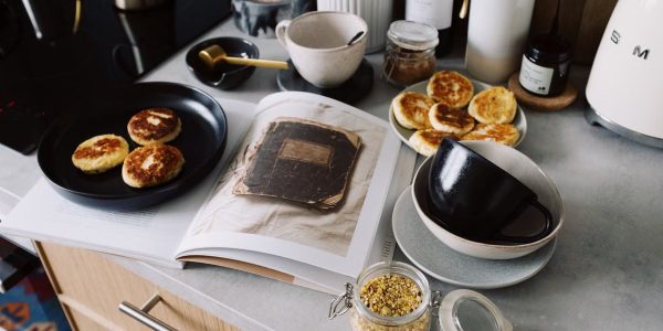 High angle of cookbook and cooked fritters on plates placed on table near utensils and ingredients in cozy kitchen at home