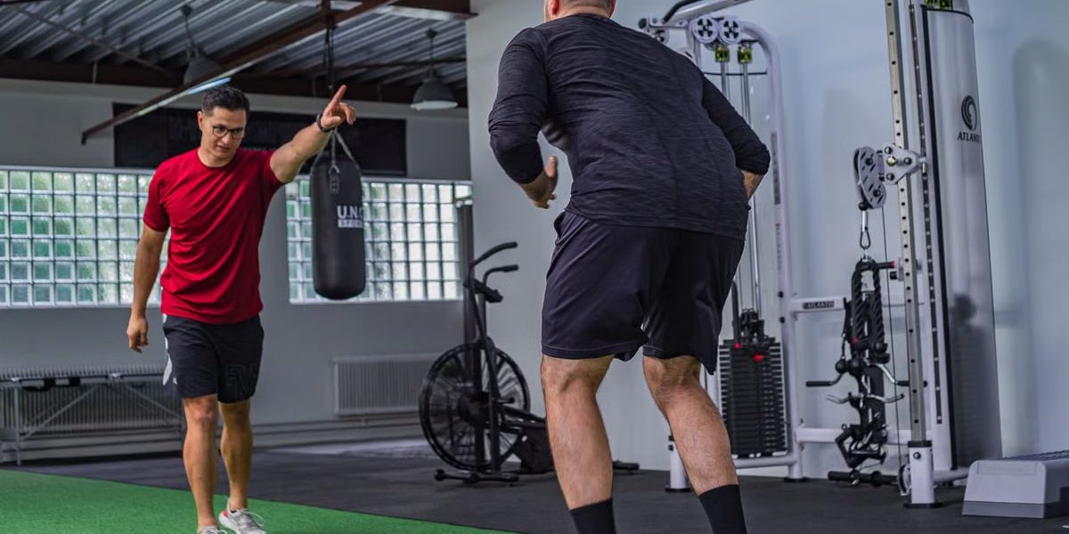 Back view of muscular man in sportswear doing exercises with trainer in modern fitness club